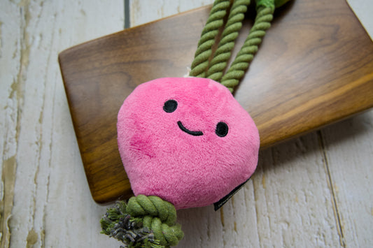 A pink plush dog toy shaped like a smiling radish with green rope handles and a knotted rope tail, displayed on a wooden board.|Un jouet en peluche rose pour chien en forme de radis souriant, doté de poignées en corde verte et d'une queue en corde nouée, présenté sur une planche en bois.