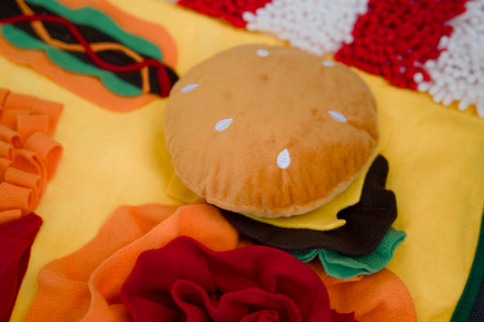 Plush toy resembling a hamburger on a colorful fabric background.|Peluche ressemblant à un hamburger sur un fond en tissu coloré.