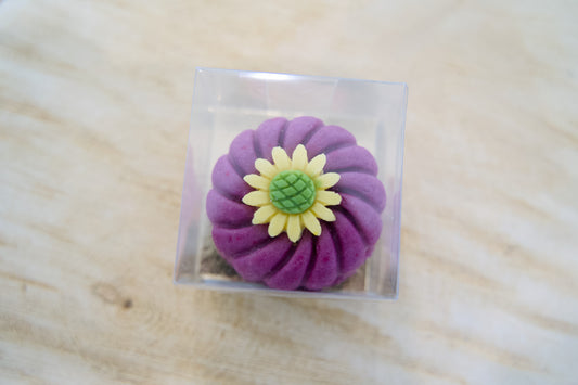 Purple flower-shaped dog cake on a clear square base with a wooden surface background.|Gâteau pour chien en forme de fleur violette, posé sur une base carrée transparente, avec un fond en bois.