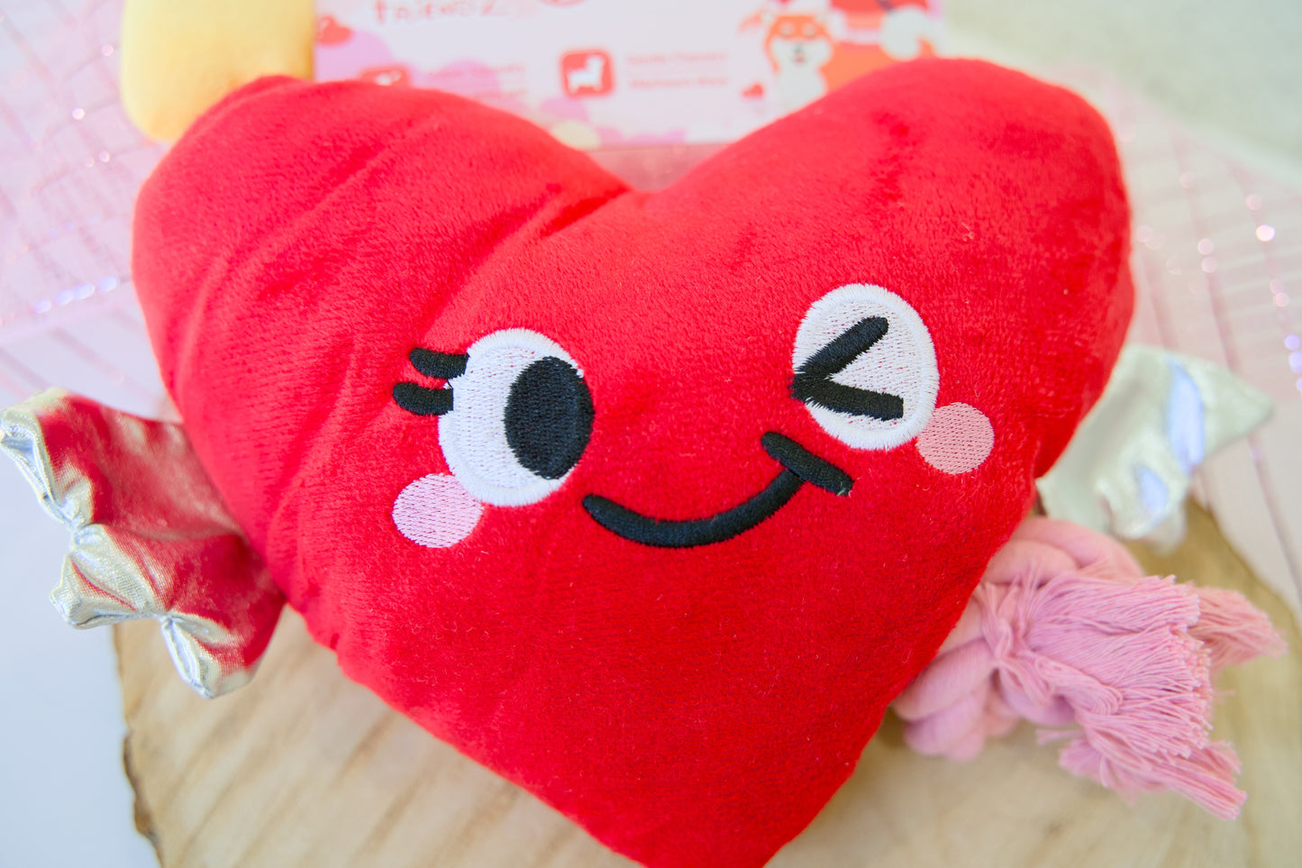 Red heart-shaped plush toy for dogs with a smiling face on a light pink background.