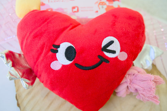 Red heart-shaped plush toy for dogs with a smiling face on a light pink background.|Peluche rouge en forme de cœur pour chiens, avec un visage souriant, sur un fond rose clair.