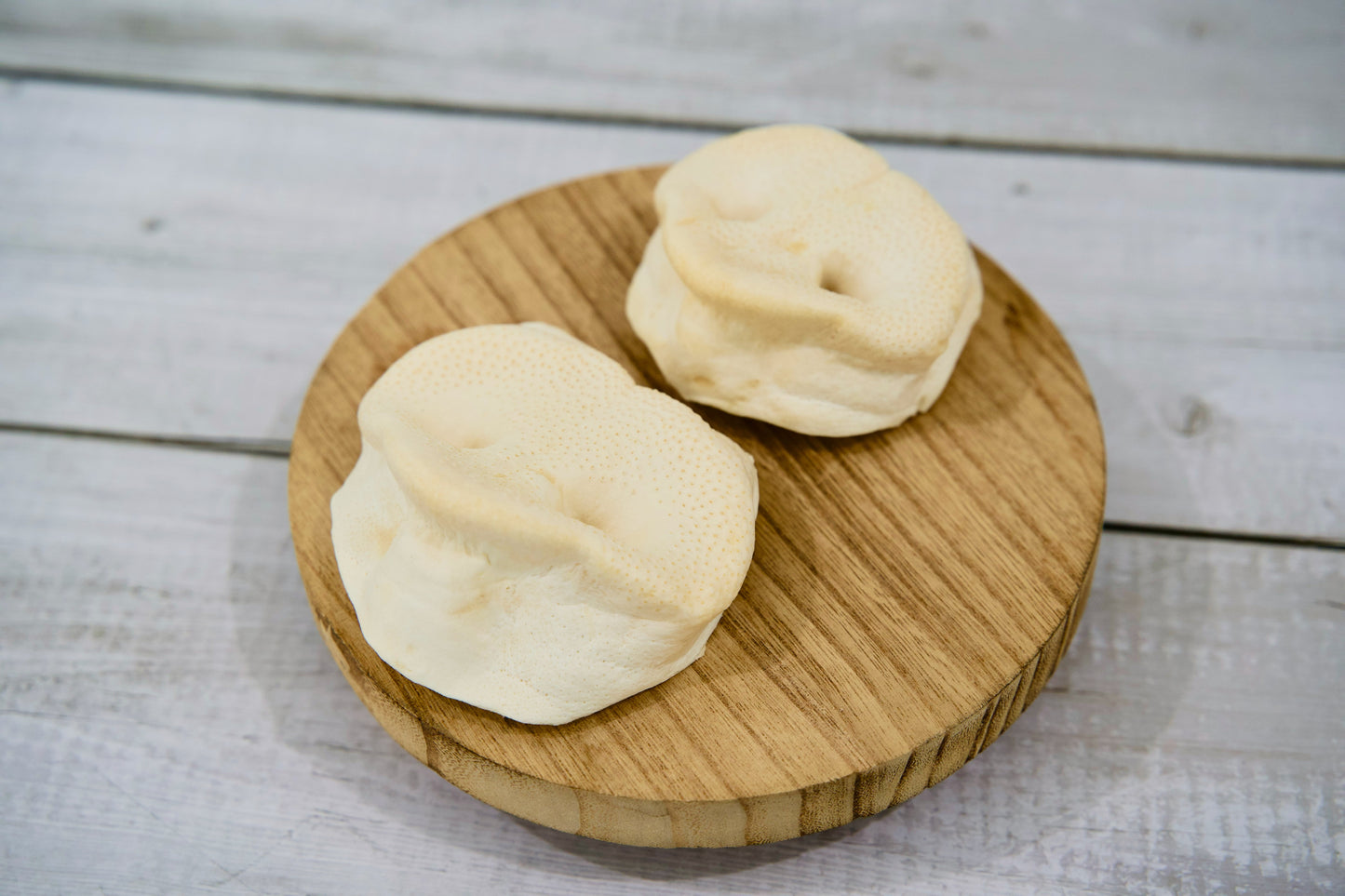 Deux friandises naturelles à mâcher pour chiens, à base de museaux de porc soufflés, présentées sur une planche ronde en bois, mettant en valeur leur texture croustillante et leur aspect entièrement naturel.