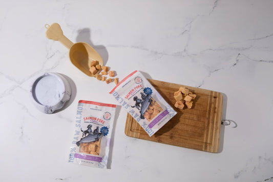 Two packages of salmon-flavored dog and cat treats are displayed on a white marble surface.|Deux paquets de friandises au saumon pour chiens et chats sont disposés sur une surface en marbre blanc.