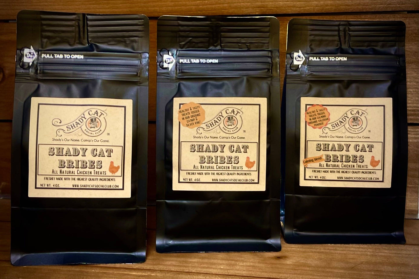 Trois paquets de friandises pour chats Shady Cat Bribes posés sur une surface en bois.