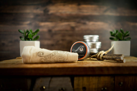 Tin of silvervine powder and towel on a wooden surface with plants and wooden background.|Une boîte de poudre de vigne argentée et une serviette posées sur une surface en bois, avec des plantes et un fond en bois.