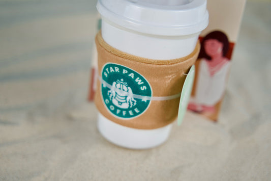 Vanilla-scented dog toy on sand, featuring rubber cup and plush sleeve for summer fun.|Jouet pour chien parfumé à la vanille sur le sable, avec tasse en caoutchouc et housse en peluche pour des jeux estivaux.