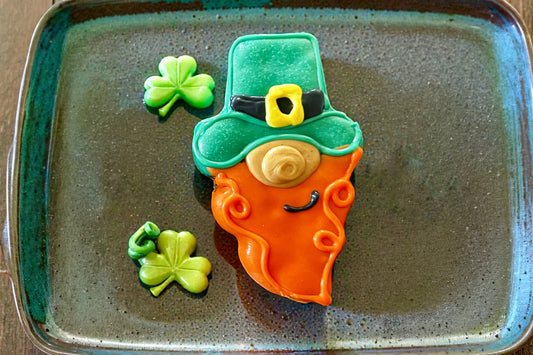 Leprechaun-shaped dog cookie with a green hat on a textured plate.|Biscuit pour chien en forme de lutin avec un chapeau vert, présenté sur une assiette texturée.