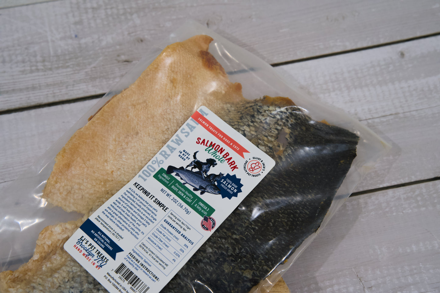 Une friandise pour chiens et chats à base de filet de peau de saumon, présentée dans son emballage sur un fond en bois blanc.