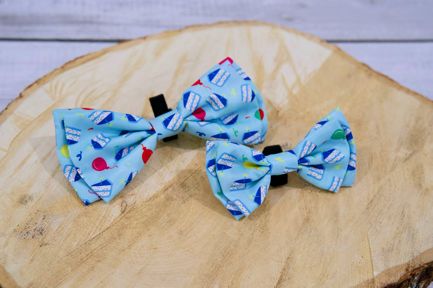 Deux nœuds papillon bleus pour animaux, ornés de motifs d'anniversaire colorés, posés sur une surface en bois.