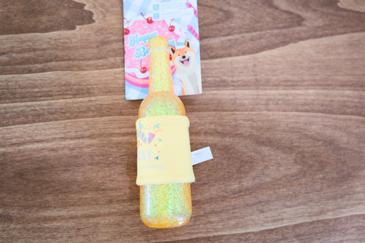 Colorful glittery bottle-shaped toy on a wooden surface with a colorful dog toy in the background.|Un jouet coloré et scintillant en forme de bouteille est posé sur une surface en bois, avec un jouet pour chien coloré en arrière-plan.