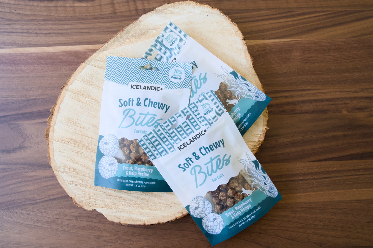 Three bags of Icelandíc+ Soft & Chewy Bites for cats on a round wood board.