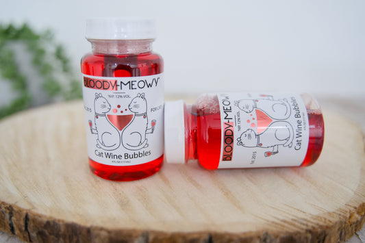 Two bottles of Bloody-Meowy cat wine bubbles on a wooden surface with a blurred background.|Deux bouteilles de vin Bloody-Meowy pour chats posées sur une surface en bois, avec un arrière-plan flou.