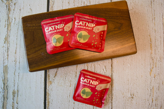 Three red packets of Catnip Flavour Dust are arranged on a wooden cutting board.|Trois sachets rouges de poudre aromatisée à l'herbe à chat sont disposés sur une planche à découper en bois.