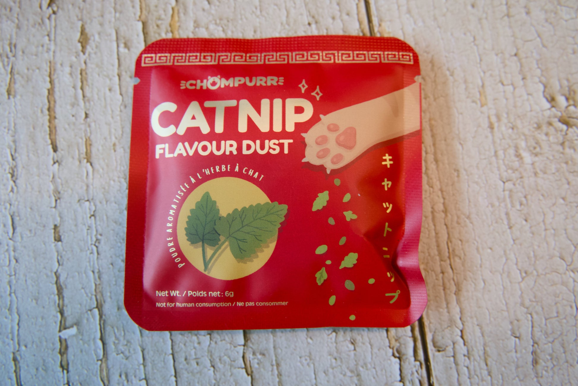 Un sachet rouge de friandises pour chats Chompurr à la poudre saveur cataire, posé sur une surface en bois blanc.
