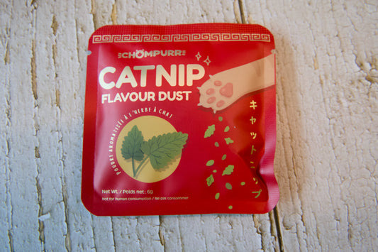 A red packet of Chompurr Catnip Flavour Dust cat treats placed on a white wooden surface.|Un sachet rouge de friandises pour chats Chompurr à la poudre saveur cataire, posé sur une surface en bois blanc.