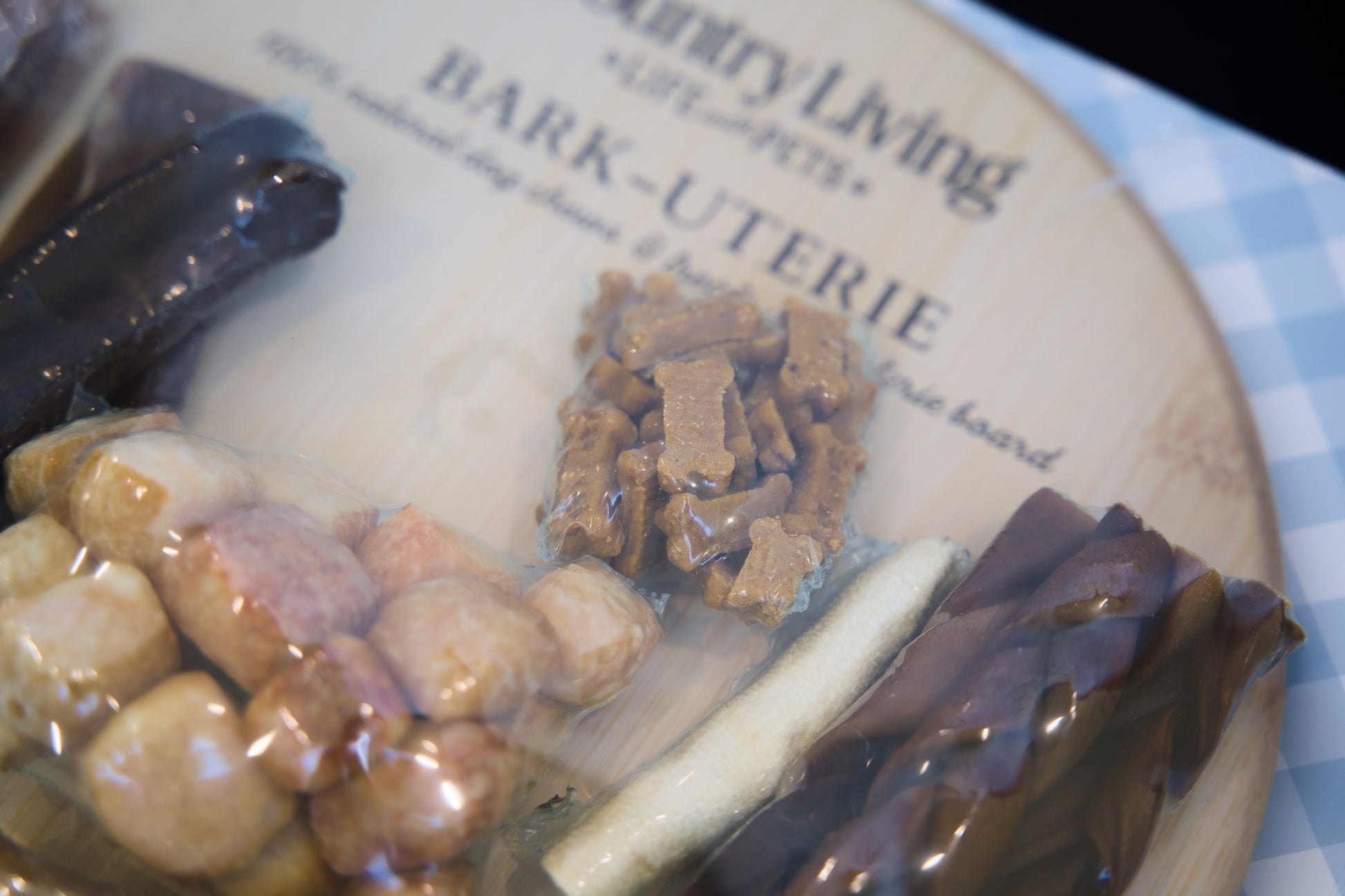 Assorted dog treats on a branded wooden board with a checkered tablecloth.