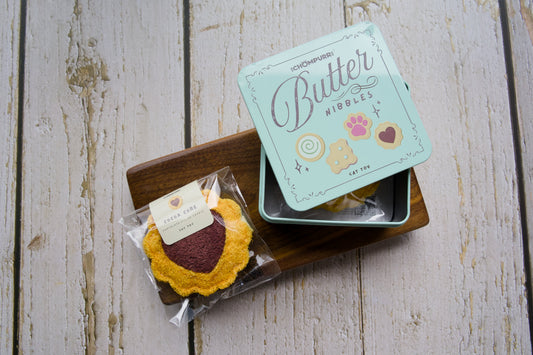 A cat toy cookie heart-shaped on a wooden tray next to its mint green tin box labeled Butter Nibbles.|Un jouet pour chat en forme de biscuit en coeur sur un plateau en bois à côté de sa boîte en métal vert menthe portant l'inscription Butter Nibbles.