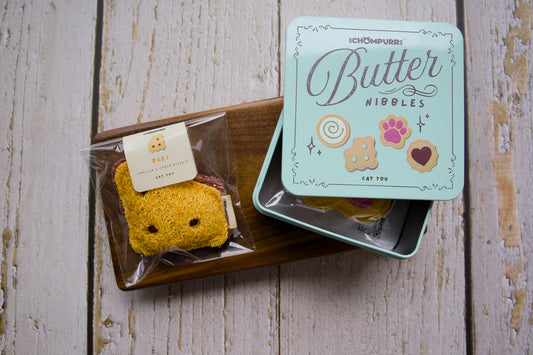 A cat toy duet cookie on a wooden tray next to its mint green tin box labeled Butter Nibbles.|Un jouet pour chat en forme de biscuit duo sur un plateau en bois à côté de sa boîte en métal vert menthe portant l'inscription Butter Nibbles.