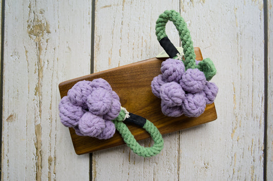 Two woven cotton grape clusters with green rope stems for dogs on a wooden board.|Deux grappes de raisin en coton tressé avec des tiges vertes en corde pour chiens, présentées sur une planche en bois.