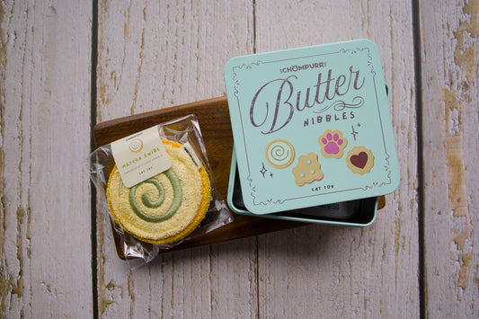 A chew cat toy filled with silvervine shaped like a matcha swirl cookie with a tin butter nibbles when buying 2 cookies.|Un jouet à mâcher pour chat garni de matatabi, en forme de biscuit roulé au matcha et une boîte en métal de biscuits au beurre offerte à l'achat de deux biscuits.