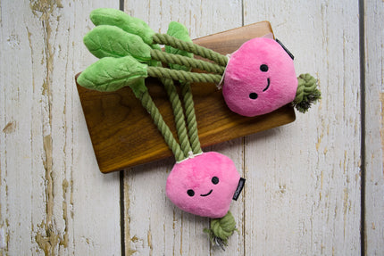 Deux jouets pour chiens roses en peluche en forme de carotte, avec des feuilles vertes et des queues en corde, présentés sur une planche en bois.
