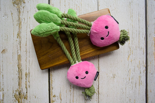Two pink plush carrot-shaped dog toys with green leaves and rope tails, displayed on a wooden board.|Deux jouets pour chiens roses en peluche en forme de carotte, avec des feuilles vertes et des queues en corde, présentés sur une planche en bois.