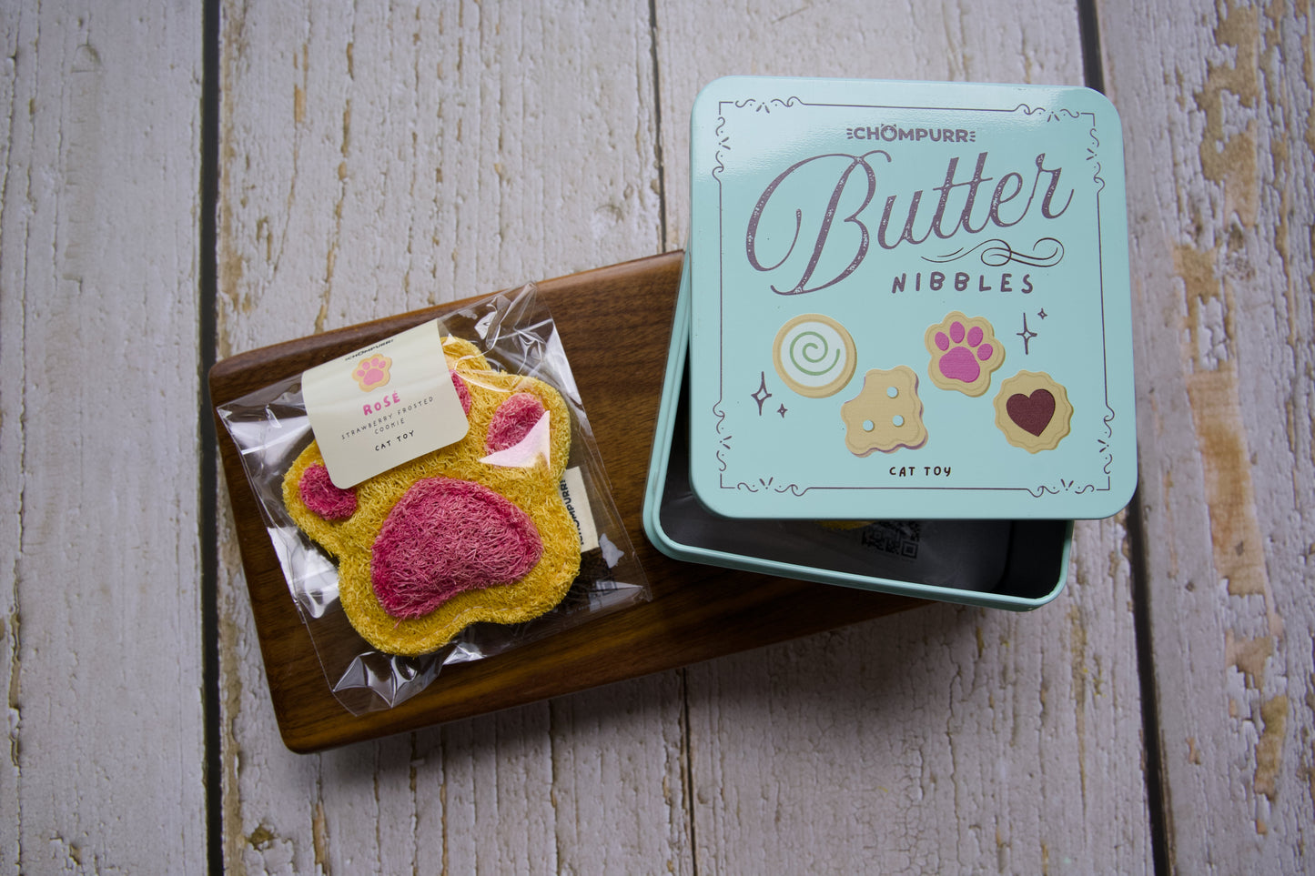 A cat toy cookie shaped like a paw print sits on a wooden tray next to its mint green tin box labeled Butter Nibbles.
