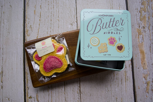 A cat toy cookie shaped like a paw print sits on a wooden tray next to its mint green tin box labeled Butter Nibbles.|Un jouet pour chat en forme de biscuit d'empreinte de patte, repose sur un plateau en bois à côté de sa boîte en métal vert menthe portant l'inscription Butter Nibbles.