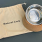 Stainless steel dog bowl with cork base and 'Natural Cork' drawstring bag on a gray background.
