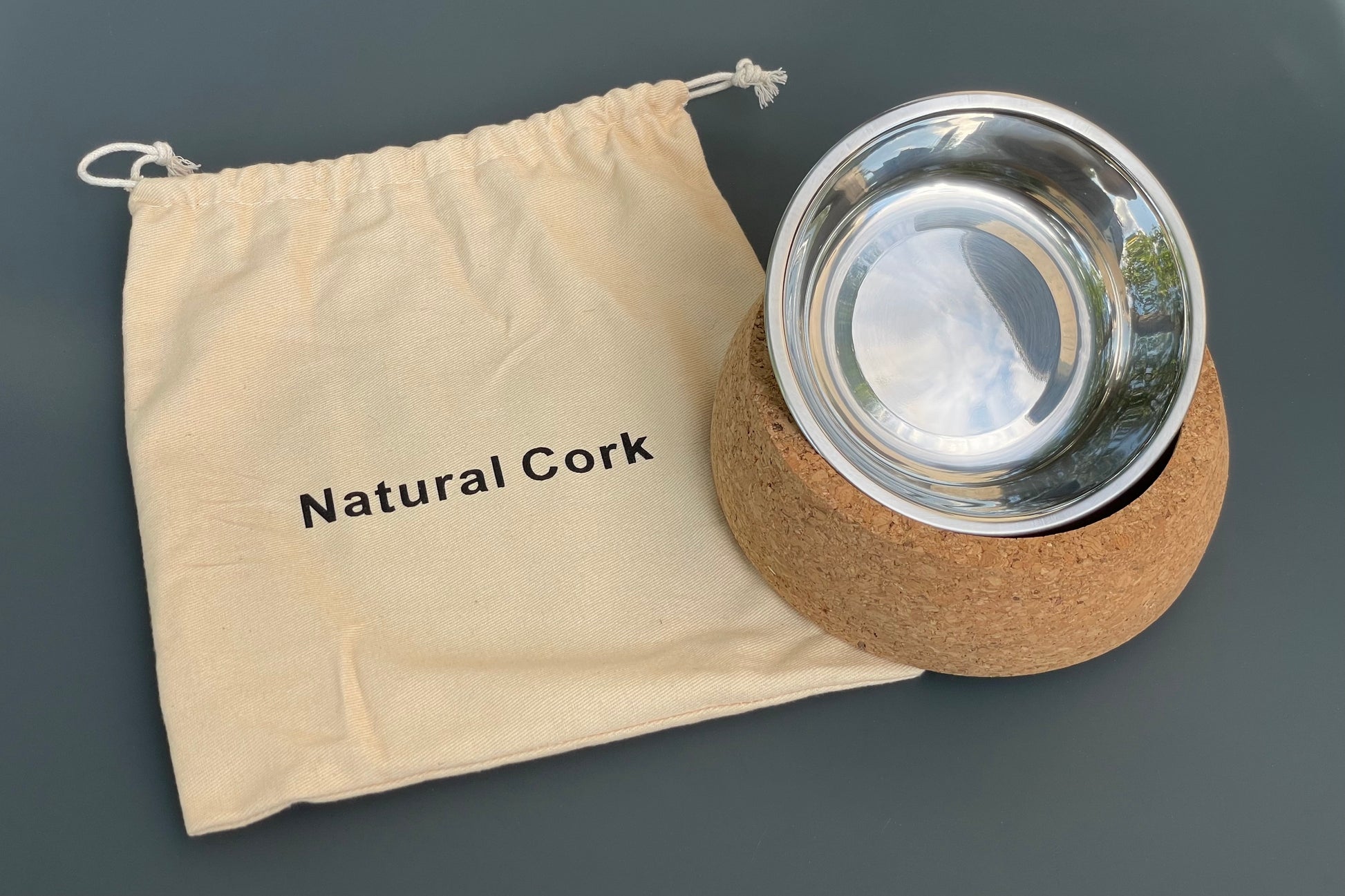 Stainless steel dog bowl with cork base and 'Natural Cork' drawstring bag on a gray background.