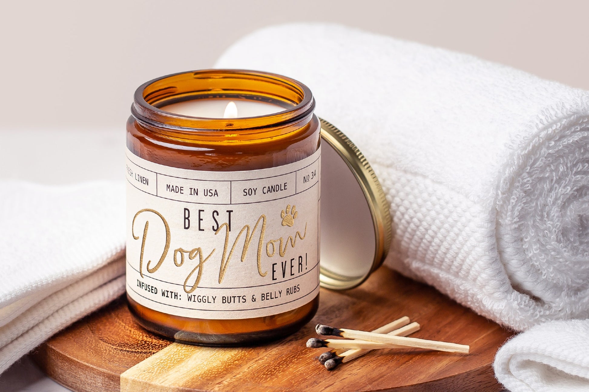 Candle in a jar labeled Best Dog Mom Ever on a wooden board with towels and matches.