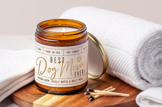 Candle in a jar labeled Best Dog Mom Ever on a wooden board with towels and matches.|Bougie dans un pot portant l'inscription Best Dog Mom Ever posée sur une planche en bois, accompagnée de serviettes et d'allumettes.