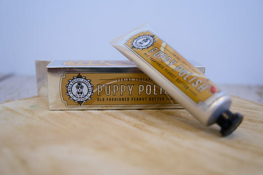 Tube of Puppy Polish peanut butter pet toothpaste on a wooden surface with a blurred background.|Tube de dentifrice pour animaux au beurre de cacahuète Puppy Polish posé sur une surface en bois avec un arrière-plan flou.