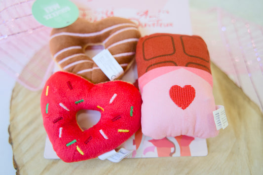 Three plush dog toys shaped like a heart with sprinkles and a chocolate bar on a wooden surface.|Trois jouets en peluche pour chiens, en forme de cœur avec des paillettes et une barre de chocolat, posés sur une surface en bois.