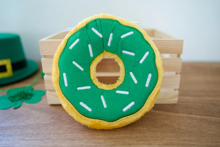 Green and yellow donut-shaped plush dog toy with a St. Patrick's Day hat in the background.