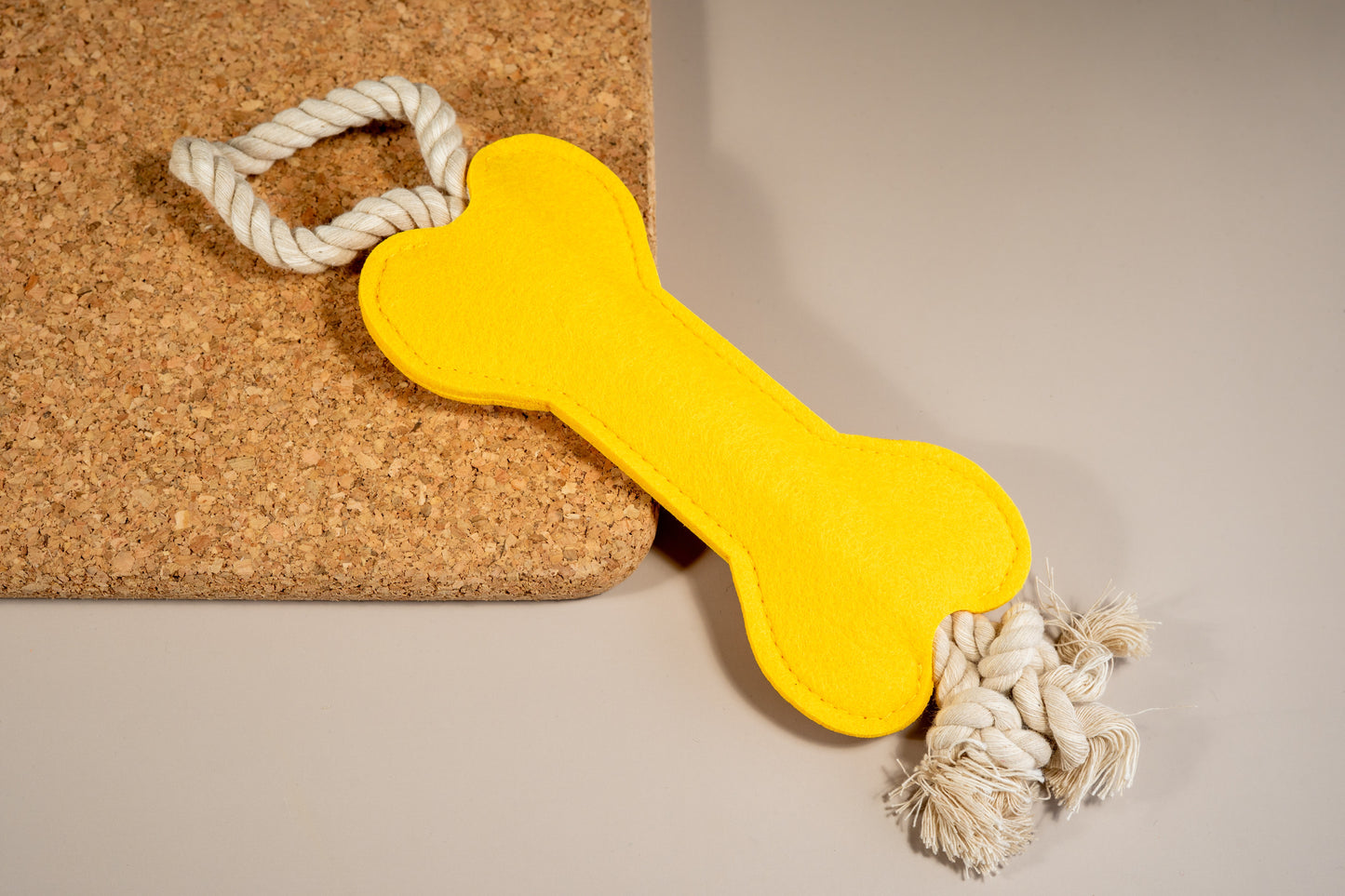 Bright yellow felt bone dog toy with rope at the ends placed on cork board.