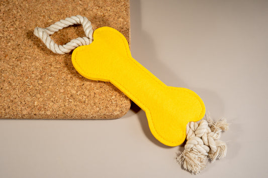 Bright yellow felt bone dog toy with rope at the ends placed on cork board. | Jouet pour chien en feutre jaune vif avec corde aux extrémités placée sur un panneau de liège.