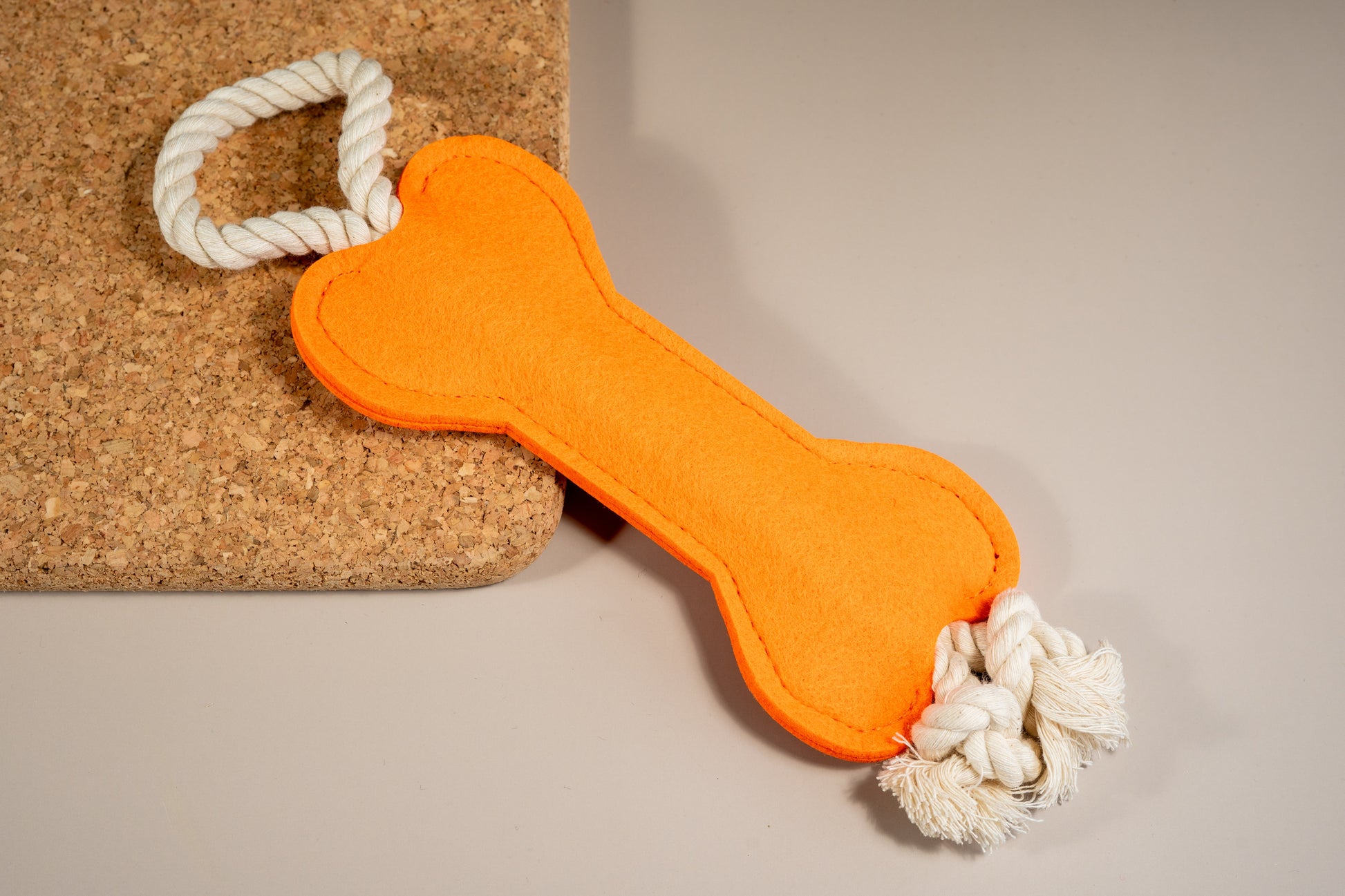 Bright orange felt bone dog toy with rope at the ends placed on cork board. | Jouet pour chien en feutre orange vif avec corde aux extrémités placée sur un panneau de liège.