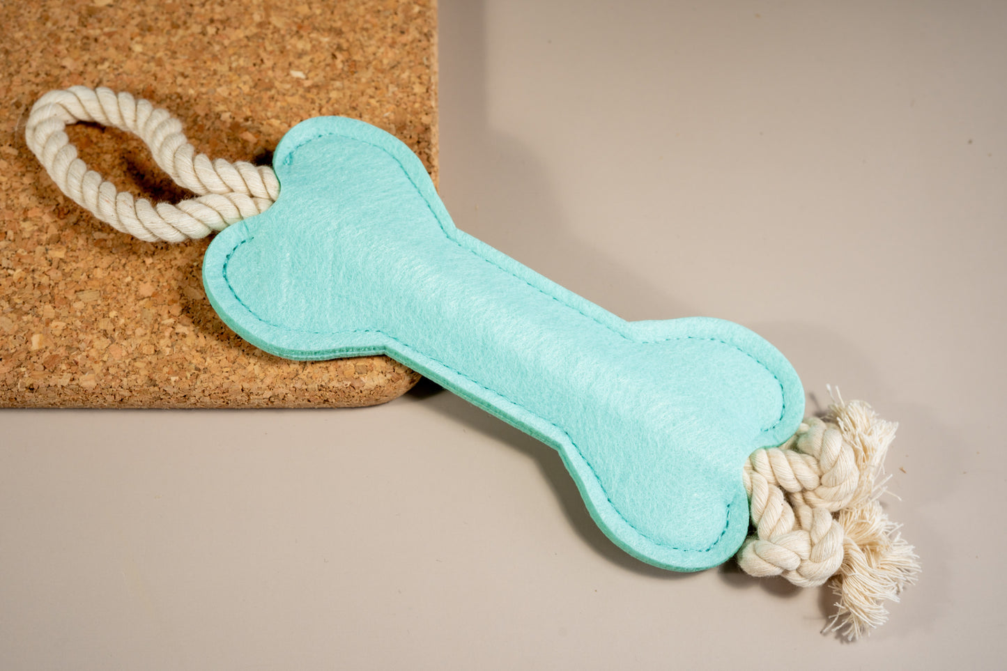 Powder blue felt bone dog toy with rope at the ends placed on cork board.