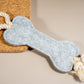 Pale gray felt bone dog toy with rope at the ends placed on a cork board.