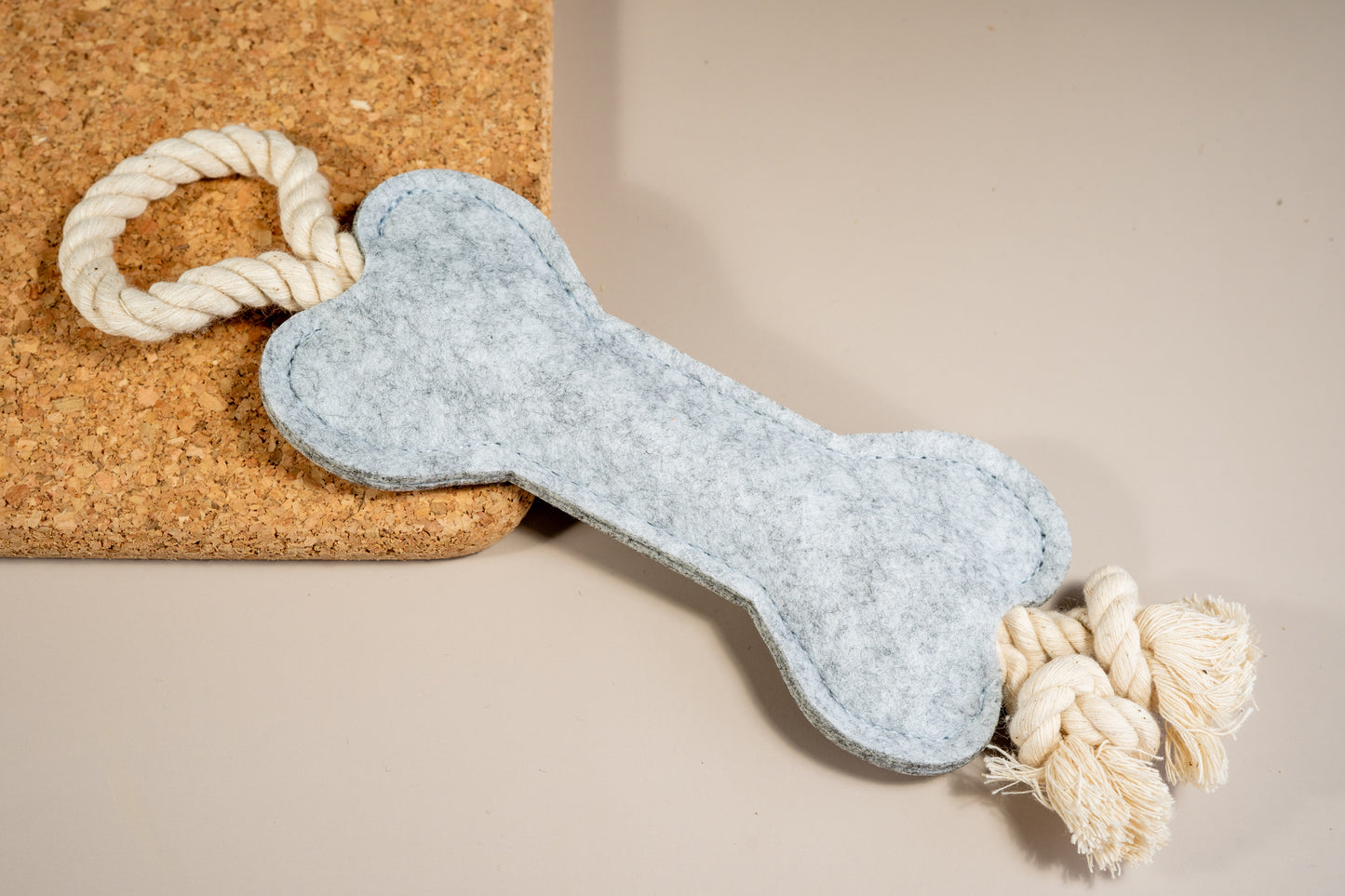 Pale gray felt bone dog toy with rope at the ends placed on a cork board.