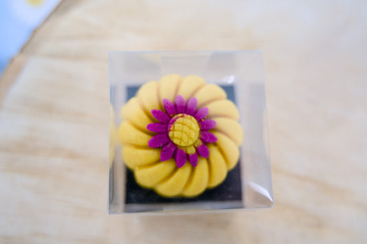 Yellow flower-shaped dog treat with a purple center in a clear plastic packaging on a beige surface.|Friandise pour chien en forme de fleur jaune avec un centre violet, présentée dans un emballage en plastique transparent sur une surface beige.