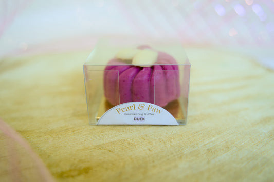 Pink duck flavour dog treat in a clear box on a yellow fabric background.|Friandise pour chien au goût de canard rose, présentée dans une boîte transparente sur un fond de tissu jaune.