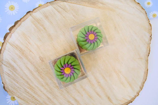 Green and purple flower-shaped dog cakes on a wooden surface with a floral background.|Des gâteaux pour chiens en forme de fleurs vertes et violettes, posés sur une surface en bois avec un fond floral.