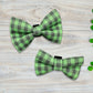Two green and black checkered dog bow ties on a wooden surface with clover leaves.