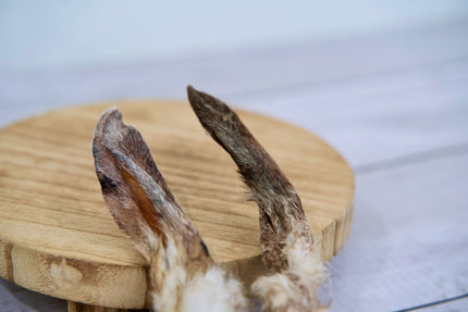 Deux friandises pour chiens à base d'oreilles de lapin séchées avec leur fourrure naturelle, présentées sur une planche en bois ronde.