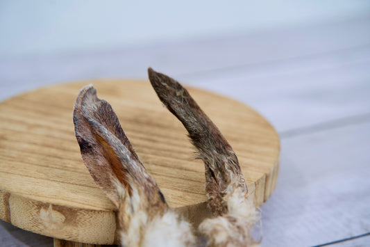 Two dried rabbit ears dog treats with natural fur, displayed on a round wooden board.|Deux friandises pour chiens à base d'oreilles de lapin séchées avec leur fourrure naturelle, présentées sur une planche en bois ronde.