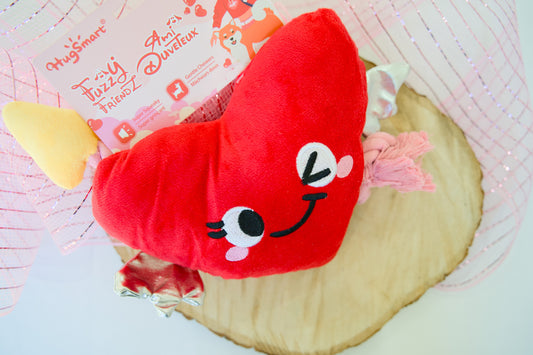 Red heart-shaped plush dog toy with a face on a wooden surface, with a pink background and text.|Jouet pour chien en peluche rouge en forme de cœur avec un visage, posé sur une surface en bois, avec un fond rose et du texte.