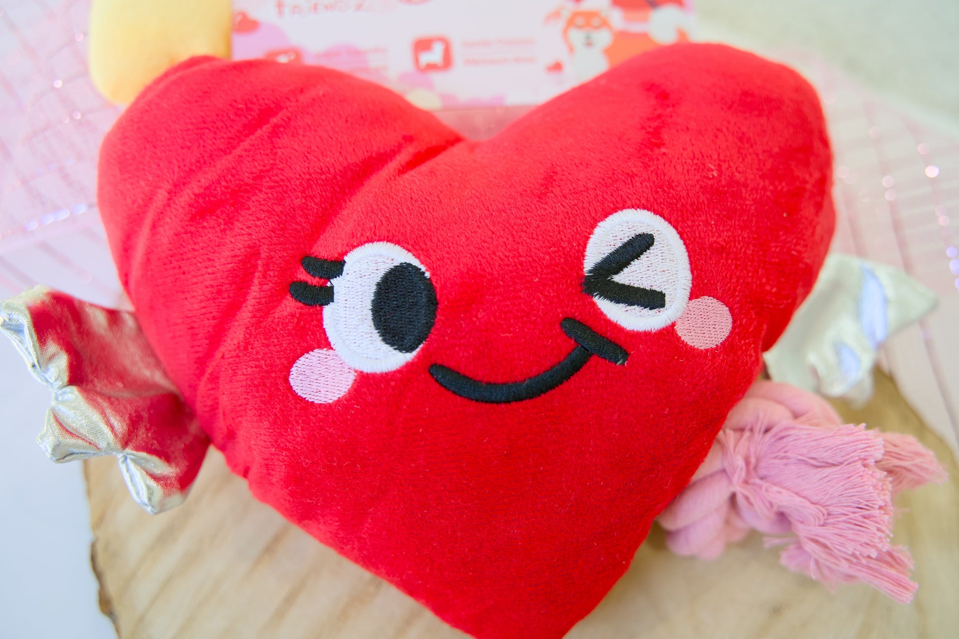 Red heart-shaped plush toy for dogs with a smiling face on a light pink background.
