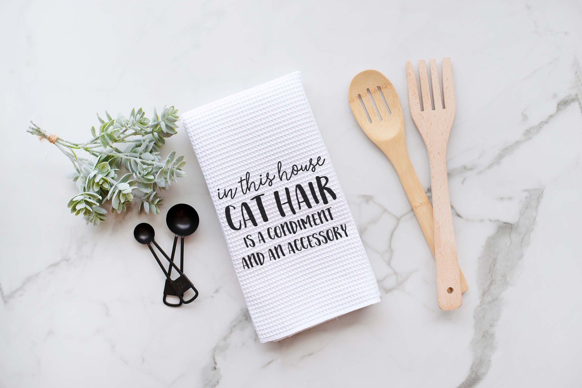 Torchon de cuisine sur le thème des chats, avec du texte, posé sur une surface en marbre aux côtés d'ustensiles en bois.